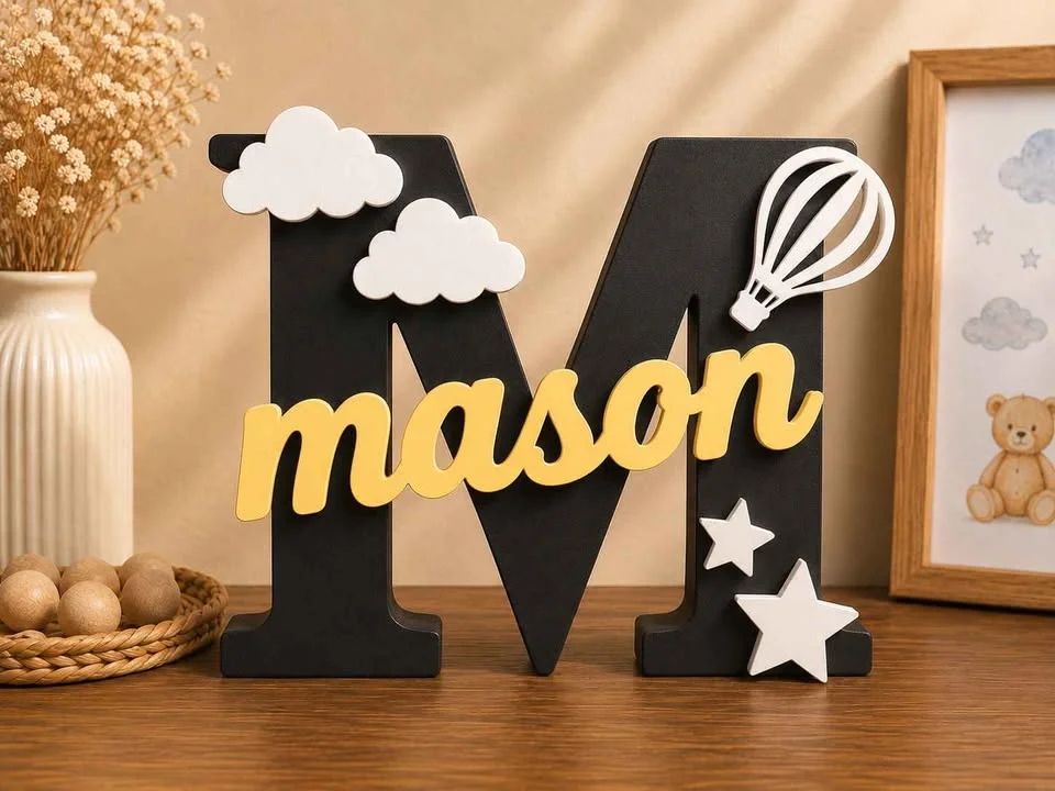 Illustration: black letter M name sign with a yellow name, clouds, a hot air balloon and stars