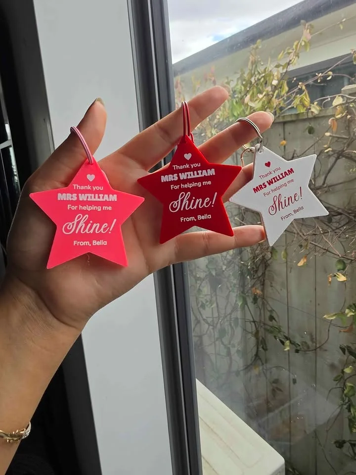 A hand holding three teacher star keychains in red, pink and white