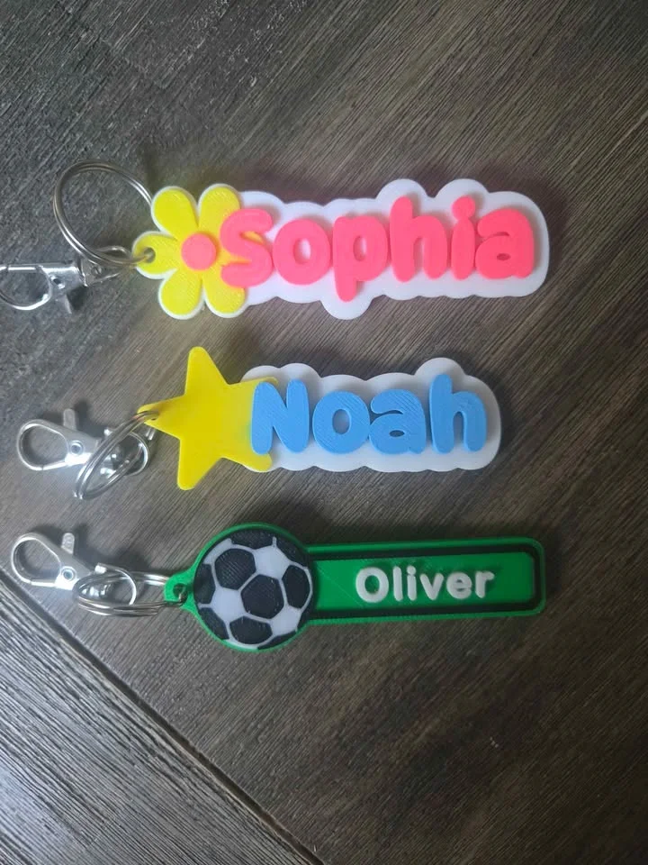Three 3D printed name bag tags: a pink name with a yellow flower, a blue name with a yellow star, and a green football design