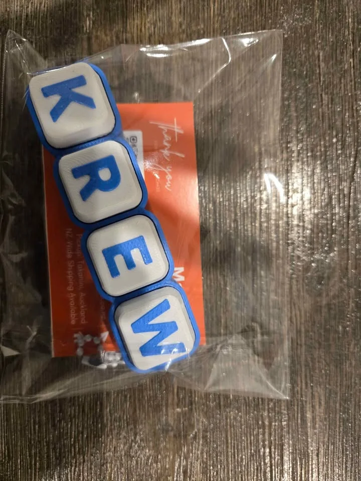 A blue clicky keyboard-key name keychain packed in a clear bag, ready to send