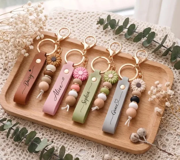 Five name keychains with daisy charms and beads in brown, pink, green, grey and blush, on a wooden tray
